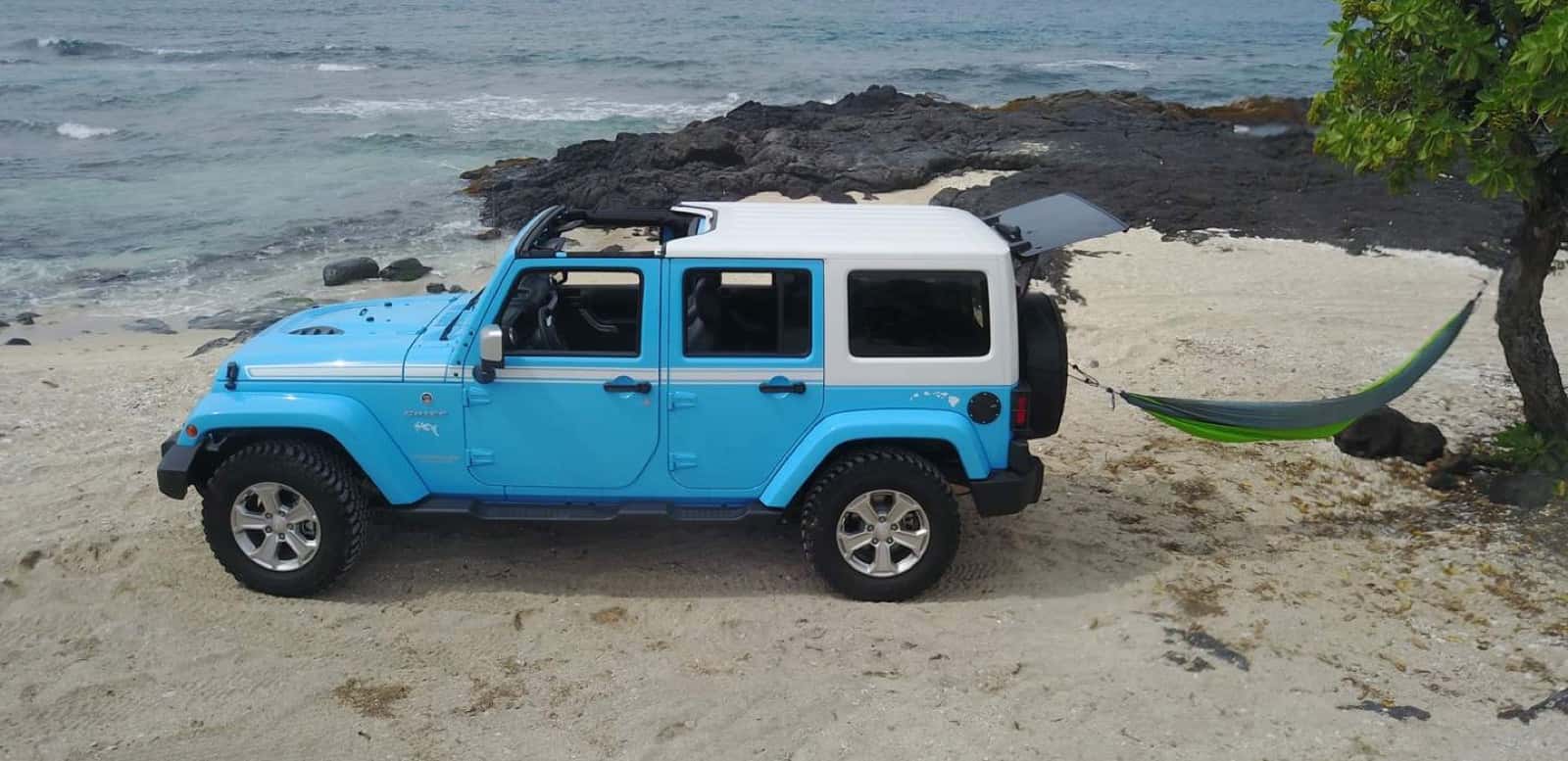 Light blue Jeep Wrangler with a white hard top on the sand beside black lava rock